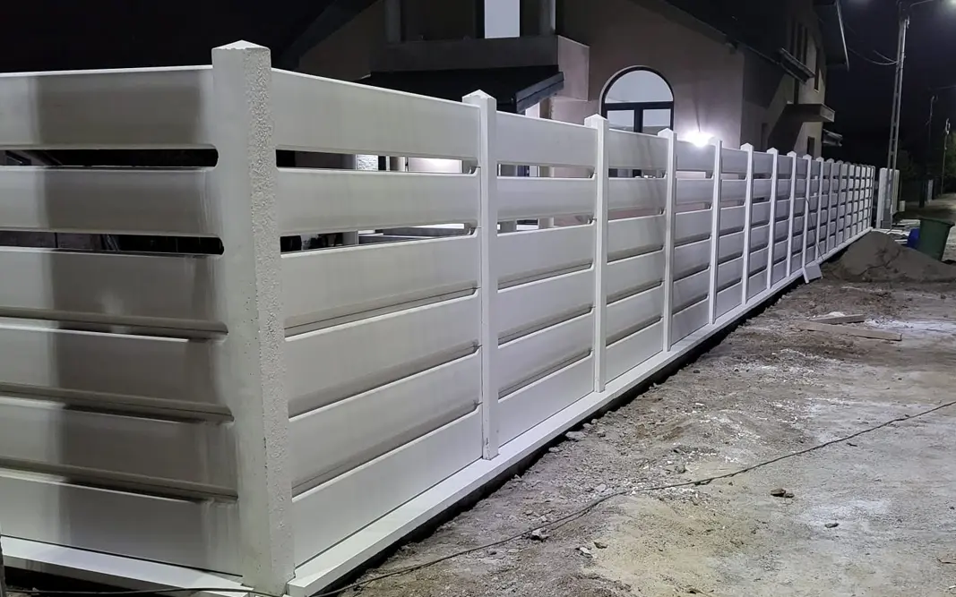Fehér betonkerítés vízszintes kerítéselemekkel egy ház előtt, esti felvétel
